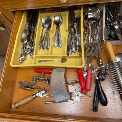Wooden kitchen drawer filled with stainless steel flatware in yellow organizers and various vintage kitchen gadgets, including can...