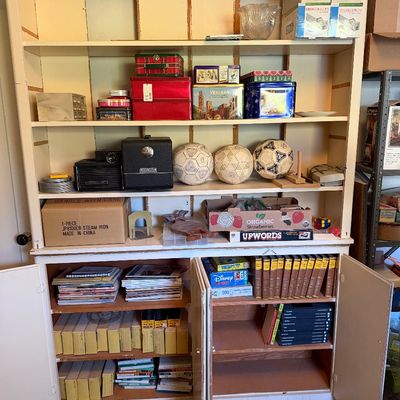 Large white wooden hutch filled with assorted vintage household goods, decorative tins, soccer balls, board games, and books.