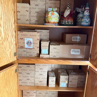 Sale Photo Thumbnail #105: A wooden cabinet filled with boxed Jim Shore Heartwood Creek collectibles, featuring decorative figurines such as a Santa Claus, pumpkin, and snowman displayed on the top shelf.