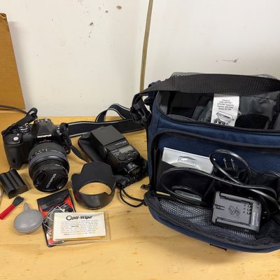 Olympus DSLR camera set including an attached lens, external flash unit, lens hood, battery charger, and cleaning supplies alongside a...