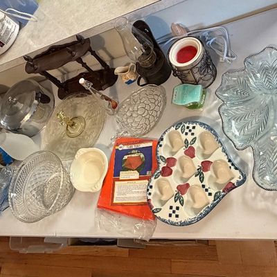 Assorted vintage household items displayed on a folding table, including cut glass serving dishes, milk glass, oil lamps, a ceramic...