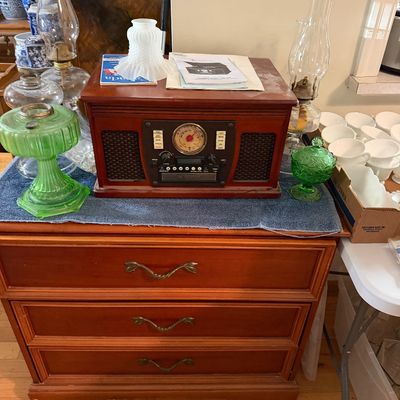A vintage-style Crosley turntable stereo console sitting on a three-drawer wooden chest, surrounded by oil lamps and green pressed glass...
