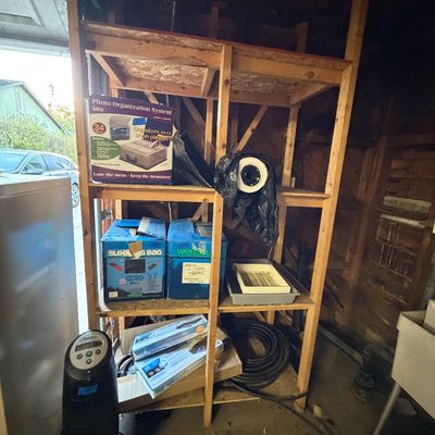 Tall wooden garage storage shelving unit holding household items, including sleeping bags in boxes, a photo organization system, and a...