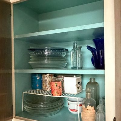 Open kitchen cabinet featuring vintage glass pie dishes, cobalt and clear glass bottles, tea tins, and a vintage poison bottle with label.