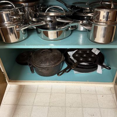 Collection of stainless steel pots and pans on the top shelf, paired with vintage cast iron skillets and a Dutch oven on the bottom shelf.