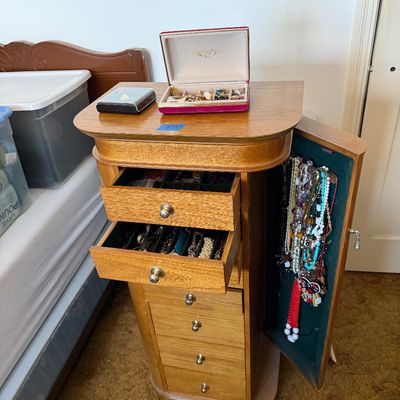 Standing wooden jewelry armoire with multiple drawers and a side cabinet filled with costume jewelry.