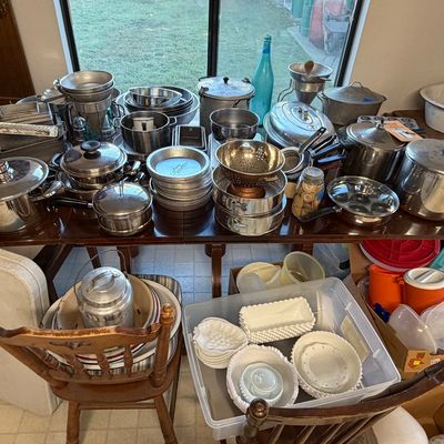 Large assortment of vintage aluminum cookware, pots, pans, colanders, and milk glass dishware displayed on tables and in storage bins.