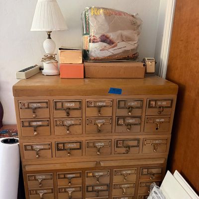 Vintage light wood multi-drawer library card catalog cabinet topped with various household goods, including a white milk glass table lamp.