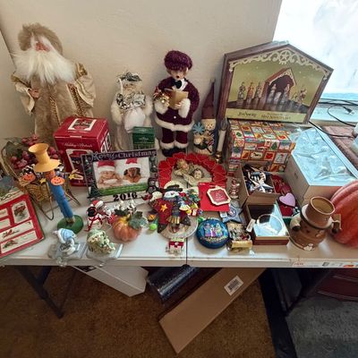 Assortment of holiday decor items arranged on a folding table, including Santa figurines, Christmas ornaments, a Jim Shore mini nativity...