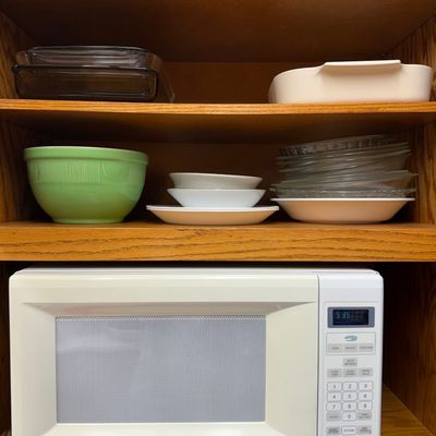 White Whirlpool countertop microwave sat below wooden shelves displaying vintage kitchenware, including a ribbed green mixing bowl, clear...