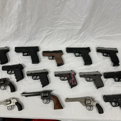 Collection of various handguns, including semi-automatic pistols and revolvers in diverse finishes, arranged on a white backdrop.