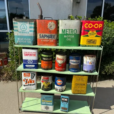 Collection of vintage oil, grease, and automotive chemical cans displayed on a light green four-tier wire shelf, including brands like...
