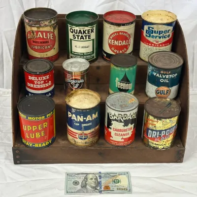 Collection of vintage oil, grease, and automotive fluid cans displayed on a wooden three-tier shelf. Cans feature classic branding...