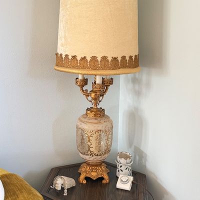 Vintage Hollywood Regency style table lamp featuring a ornate gold-toned metal and glass base with a tall fabric shade trimmed in lace.