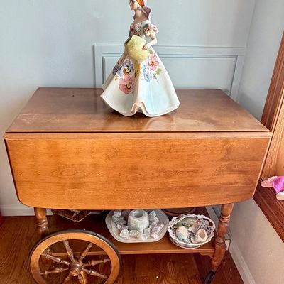 Vintage mid-century wooden drop-leaf tea cart featuring spoked wagon wheels, displayed with a hand-painted porcelain woman figurine on top.