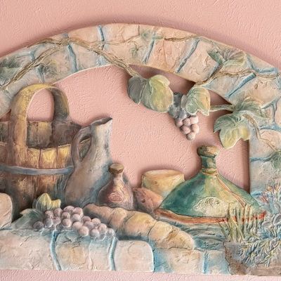 Vintage molded 3D relief wall plaque depicting a rustic Tuscan still life scene with grapes, wine, and baskets, mounted on a pink wall.