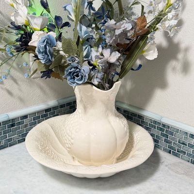 Vintage white ceramic pitcher and basin set filled with an arrangement of faux blue and white flowers.