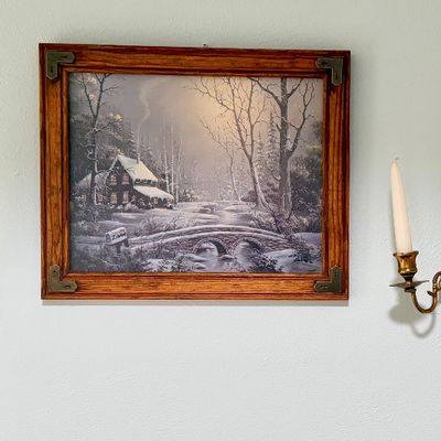 Framed Thomas Kinkade winter cottage print flanked by a pair of three-arm brass wall sconces holding taper candles.
