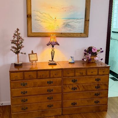 Pair of matching traditional maple or pine chest of drawers placed side by side, topped with decor and illuminated by a small table lamp...