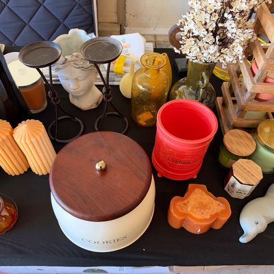 Sale Photo Thumbnail #159: Collection of home decor items featuring a vintage white cookie jar with wooden lid, jar candles, iron candlestick stands, amber glassware, and a red utensil crock.