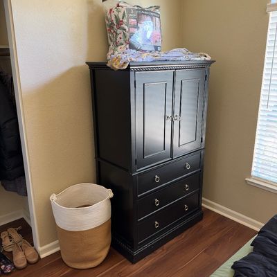 Black painted wooden armoire featuring a top cabinet and three lower drawers, paired with a two-tone woven storage basket.