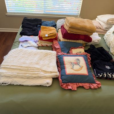 Assortment of household linens, folded towels, throw blankets, decorative pillows, and clothing laid out on a bed.
