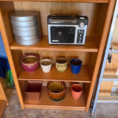 Sale Photo Thumbnail #109: Wooden bookcase filled with mixed items including metal film canisters, a vintage General Electric portable radio, and various ceramic plant pots.