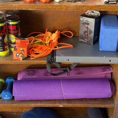 Sale Photo Thumbnail #148: Wooden shelving unit filled with exercise equipment, including purple yoga mats, dumbbells, Penn tennis balls, KT Tape, and resistance bands.