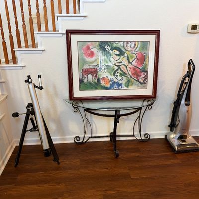 Framed Marc Chagall art print displayed on a glass-top wrought iron console table, flanked by a Bushnell telescope on a tripod and an...