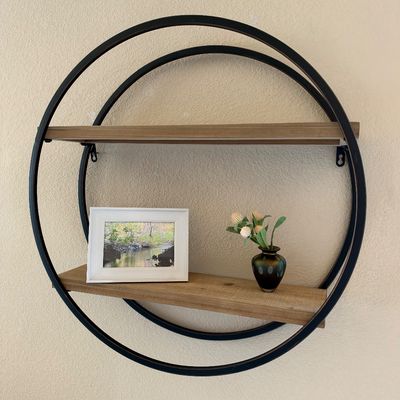 Modern circular metal frame wall shelf with two wooden display tiers, featuring a framed landscape photo and decorative small vase.