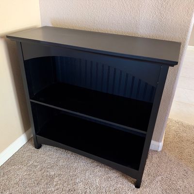 Black painted wooden low bookcase featuring two fixed shelves, an arched top apron, and beadboard backing.