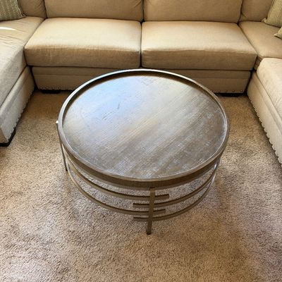 Contemporary round wooden coffee table with a tray-top style surface and a brushed metal frame, set in front of a beige sectional sofa.