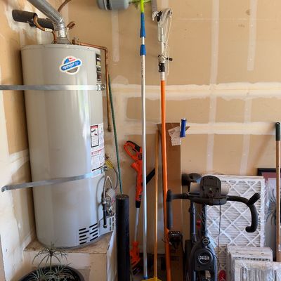 Sale Photo Thumbnail #111: Collection of garage and household equipment including a DIHASUN stationary exercise bike, long-handled cleaning tools, a pole saw, and HVAC air filters alongside a water heater.