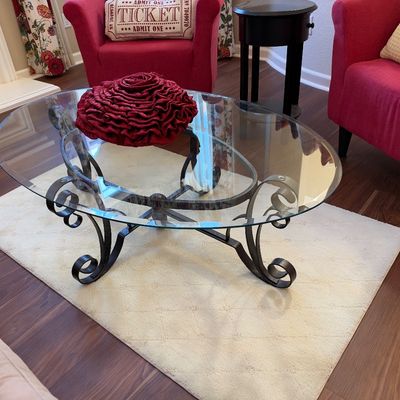 Oval glass-top coffee table featuring a wrought iron scrollwork base, paired with a small beige area rug.
