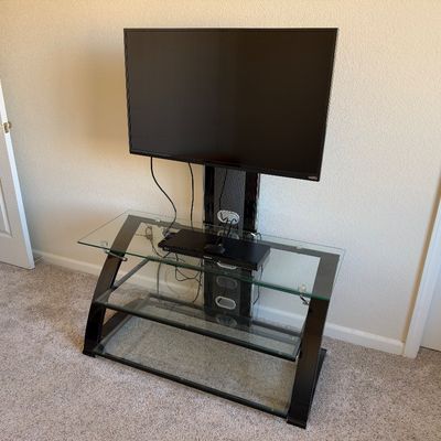 Vizio flat-screen television mounted on a contemporary glass and black metal media stand, featuring three tiers of tempered glass shelving.