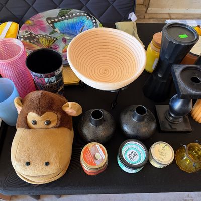 Sale Photo Thumbnail #160: Assorted household goods, including a plush monkey slipper, banneton proofing basket, black candle holders, small vases, and jar candles, displayed on a black table.