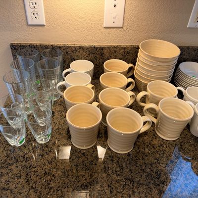 Large collection of kitchenware, featuring clear glass drinking cups alongside cream-colored ceramic mugs and stacked bowls.