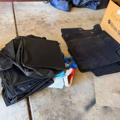 Collection of black reusable fabric shopping bags alongside a set of black carpeted vehicle floor mats.
