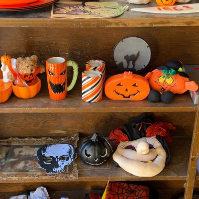 Sale Photo Thumbnail #143: Collection of vintage and modern Halloween decor, including jack-o'-lantern mugs, candles, plush figures, and masks on wooden shelves.