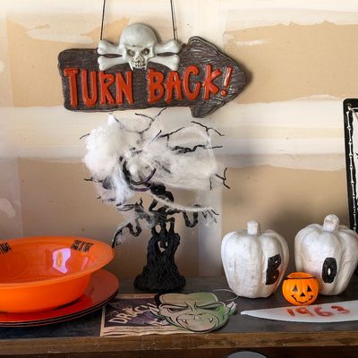 Sale Photo Thumbnail #144: Collection of Halloween decor including a hanging 'Turn Back!' skull sign, a black spooky tree centerpiece with faux cobwebs, three white painted pumpkins spelling out 'BOO', and an orange trick-or-treat bowl.