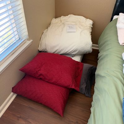 Stack of bedding and pillows on the floor, featuring a folded white mattress topper labeled "California King" alongside red...