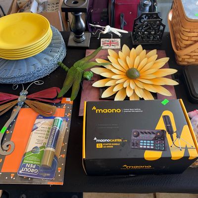 Sale Photo Thumbnail #153: Tabletop assortment of items including yellow Fiesta dinner plates, decorative metal dragonflies, a boxed Maonocaster Lite microphone kit, and a woven picnic basket.