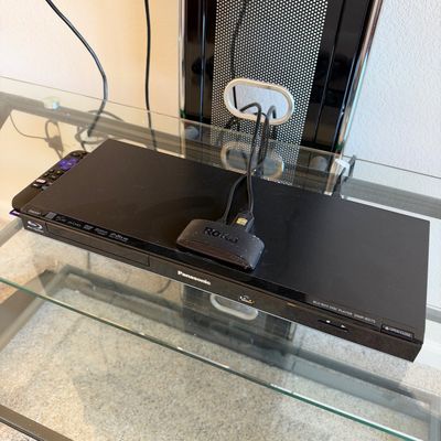 Black Panasonic DMP-BD75 Blu-ray disc player resting on a glass media stand, paired with a Roku streaming device and remote.