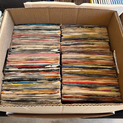 Sale Photo Thumbnail #170: Cardboard box densely filled with a large collection of vintage 45 RPM vinyl records in paper sleeves.