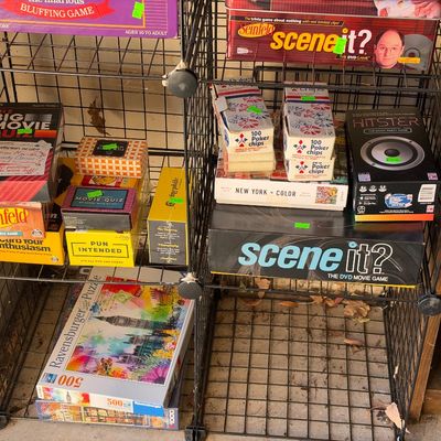 Sale Photo Thumbnail #138: Wire shelving unit filled with vintage and modern board games, trivia games, and jigsaw puzzles, including Scene It?, Seinfeld trivia, Hitster, and Ravensburger puzzles.