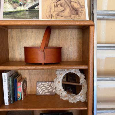 Sale Photo Thumbnail #108: Light oak bookcase housing a bentwood shaker-style carrier basket, macramé wall mirror, decorative plaque, and a selection of books.