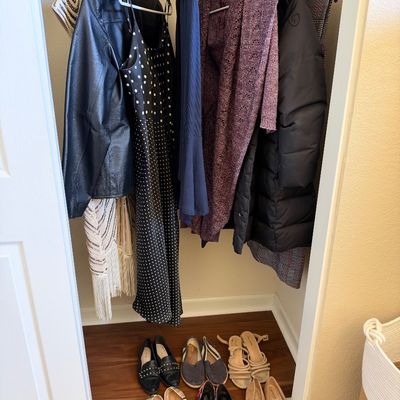 Selection of women's clothing hanging in a closet, including a leather jacket, polka dot dress, cardigan, and winter coat, alongside...