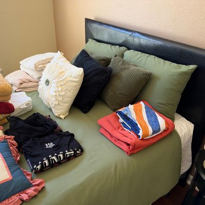 Queen-size bed with a dark faux-leather headboard, dressed in olive green linens and stacked with various throw pillows, folded towels,...