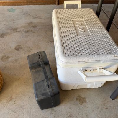 Large vintage white Igloo ice chest cooler sitting on a concrete floor next to a hard plastic carrying case and a woven basket.