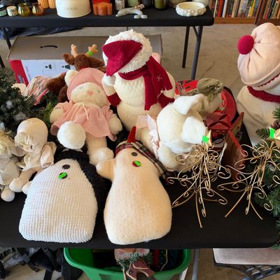 Sale Photo Thumbnail #124: Collection of holiday decor including fabric snowmen, plush dolls, mini artificial Christmas trees, and metal wire tree stands on a folding table.